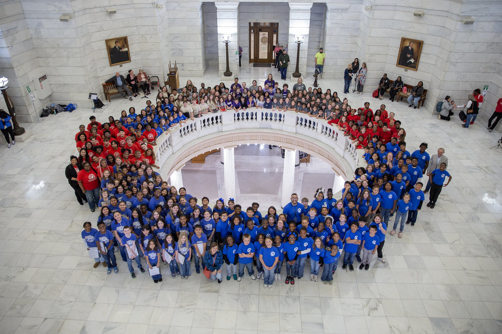 A large group of people wearing color‑coordinated shirts stand in a circular formation inside the Arkansas State Capitol building with columns and a balcony.
