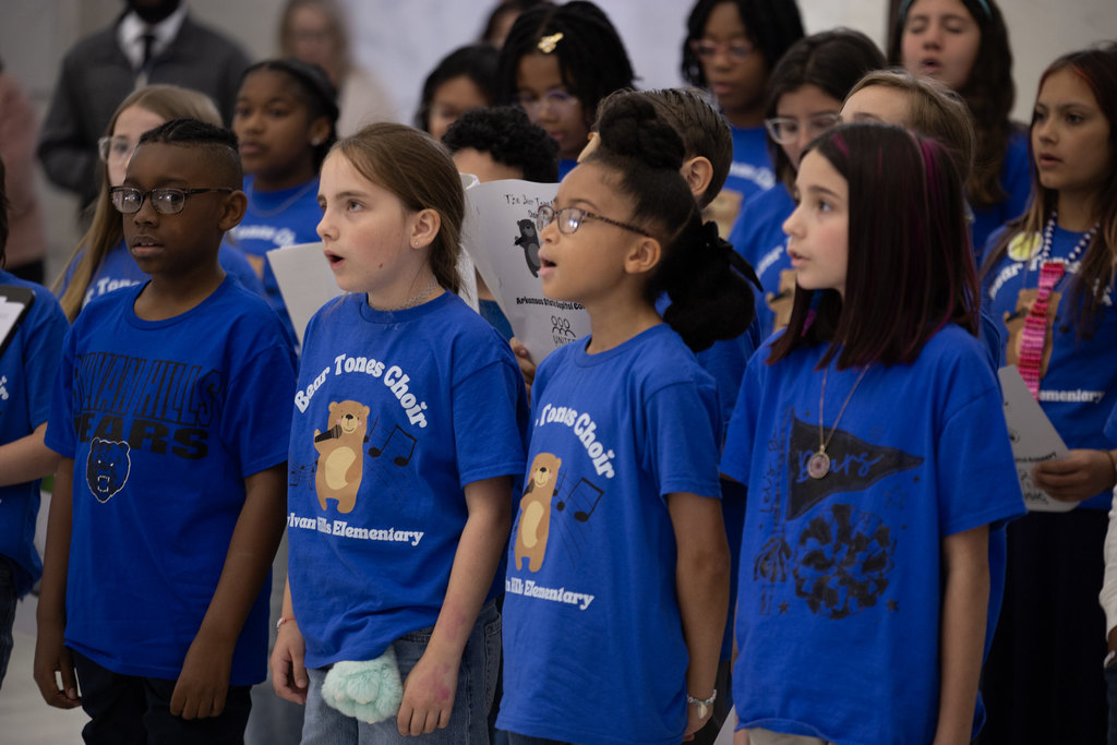 Elementary students in matching blue choir shirts sing together while holding lyric sheets. Other students and adults watch from behind them in a busy indoor space.