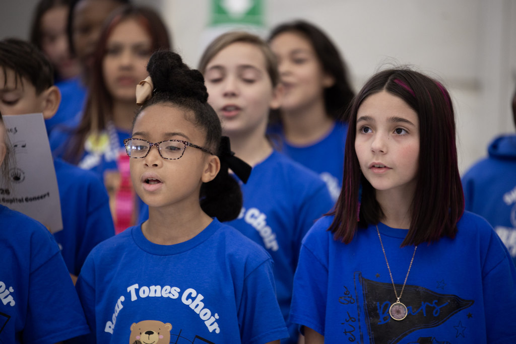 A group of children in blue “Bear Tones Choir” shirts sing while holding lyric sheets. They stand indoors with other students visible in the background.