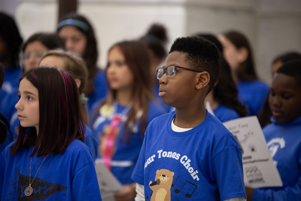A group of children in blue choir shirts sing with focused expressions. Musical note and bear graphics are visible on some shirts, and the group stands against a light-colored indoor wall.