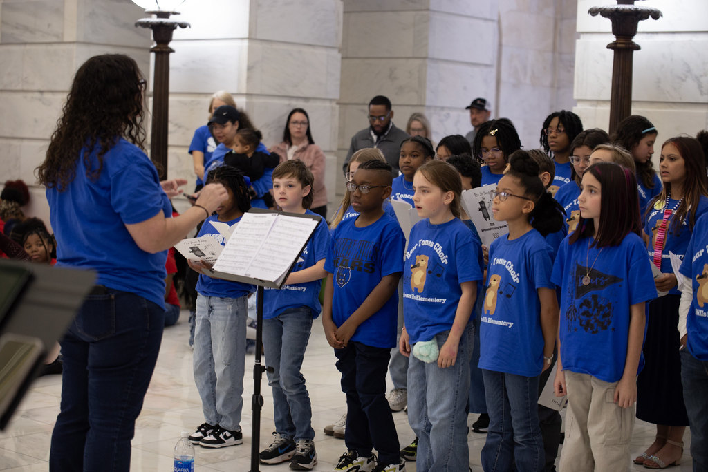 A group of elementary students in matching blue “Bear Tones Choir” shirts sing together inside a marble-walled public building. A teacher stands in front of them conducting with both hands raised, and a music stand with sheet music is positioned nearby. Observers stand in the background.