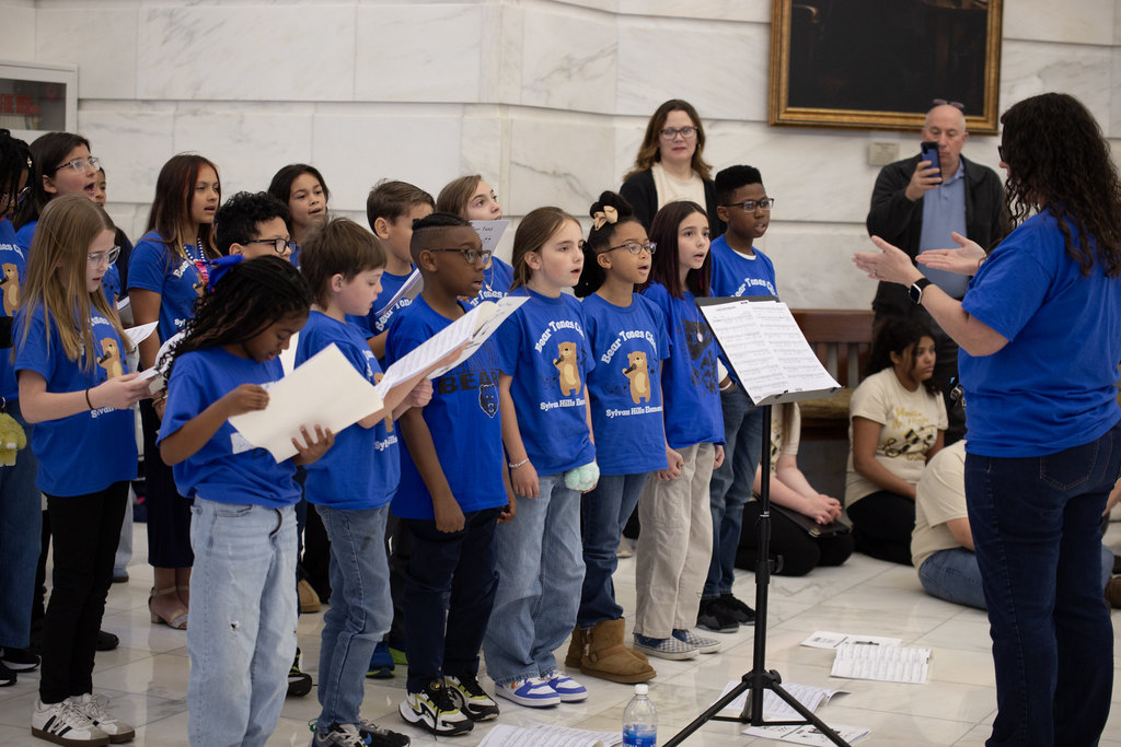 Children wearing blue “Silver Tones Choir” shirts sing together in the Arkansas State Capitol rotunda. Several hold lyric sheets.