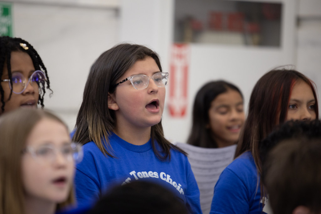 Children in matching blue “Bear Tunes Choir Sylvan Hills Elementary” shirts sing while holding lyric sheets. A teacher stands in front conducting, with a music stand between them. Adults observe from the background in a marble-walled building.
