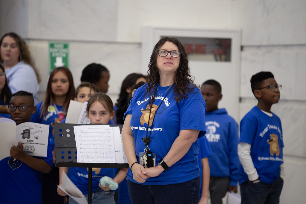 An adult and several children wearing blue “Bear Tunes Choir Sylvan Hills Elementary” shirts stand together during a performance. A music stand displays sheet music, and one child holds a booklet labeled “2024 State Capitol Concert Arkansas Music.”