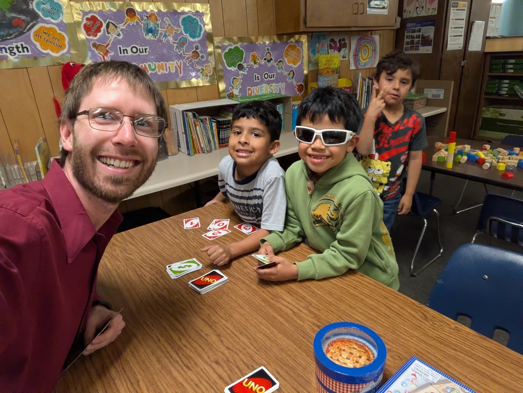 An adult in a maroon shirt sits at a classroom table playing UNO with three children. One child wears white sunglasses and a green hoodie, another wears a striped shirt, and a third stands nearby in a SpongeBob shirt. Colorful posters and learning materials fill the background.
