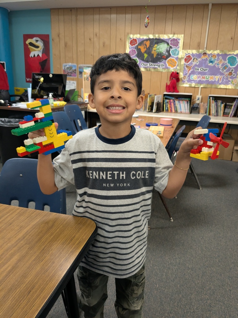 A child wearing a gray striped “Kenneth Cole New York” shirt holds up two colorful LEGO creations in a classroom decorated with posters about community and diversity. Blue chairs, bookshelves, and classroom supplies fill the background.