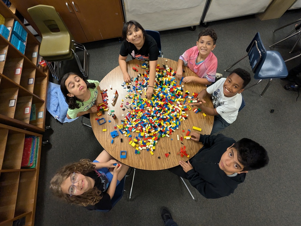 Six children sit around a circular table overflowing with colorful LEGO bricks. They look up toward the camera, smiling, with the classroom shelves and chairs visible around them. The scene captures collaborative building and creative play.