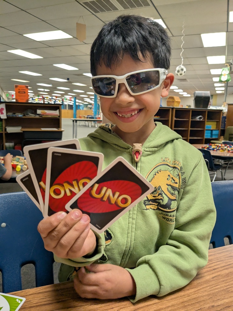 A child wearing white sunglasses and a green Jurassic World hoodie smiles while holding four UNO cards. They sit at a classroom table with shelves and decorations visible behind them.
