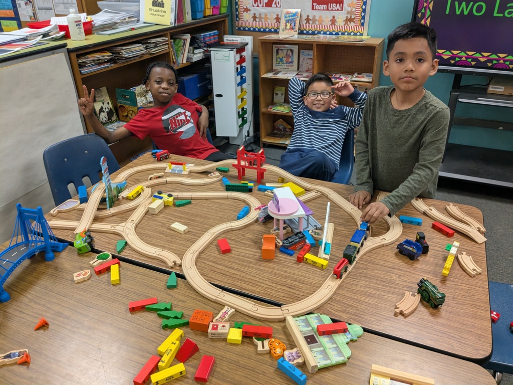 Three children gather around a classroom table covered with an elaborate wooden train‑track layout. The table is filled with toy trains, cars, bridges, and colorful blocks. Shelves, posters, and a screen in the background create a lively learning environment as students explore hands‑on building activities.