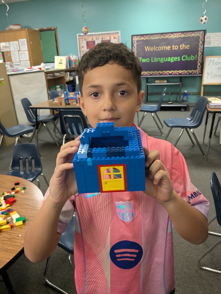 A child stands in a classroom holding a small blue LEGO structure with a red and yellow door. Behind them, a round table is scattered with LEGO pieces, and a screen displays “Welcome to the Two Languages Club!” in bright colors.