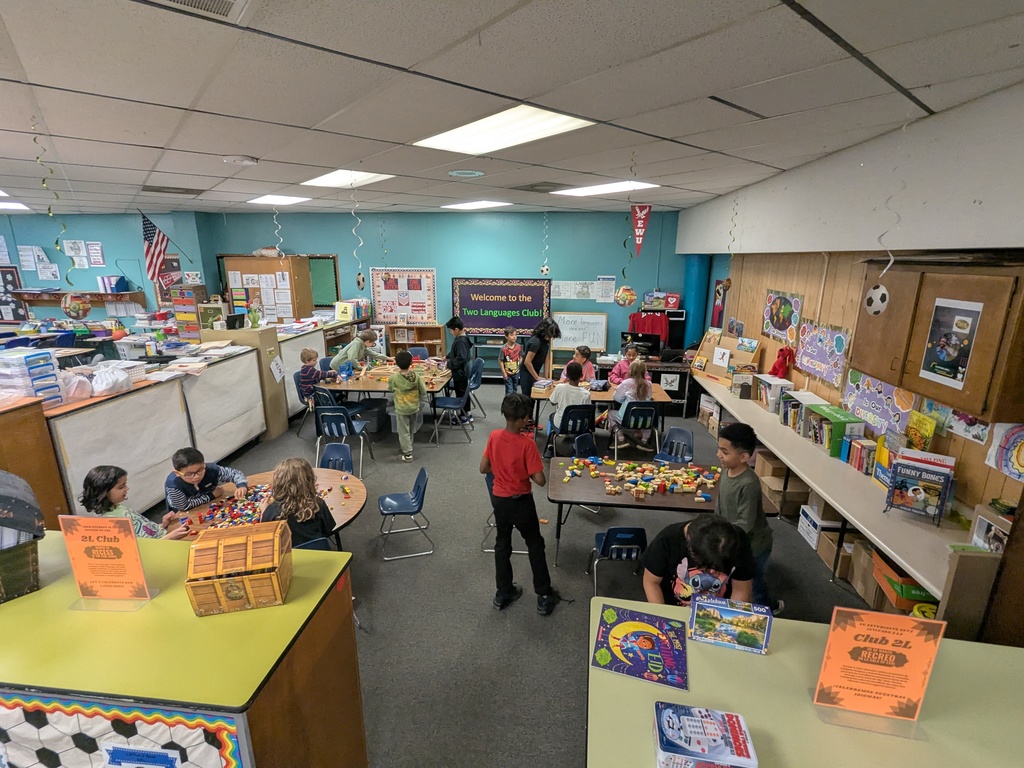 A classroom full of children works at tables on building activities with blocks and educational toys. The room is decorated with bookshelves, posters, and a bulletin board that reads “Welcome to the Two Languages Club!” and “More languages means more fun.”
