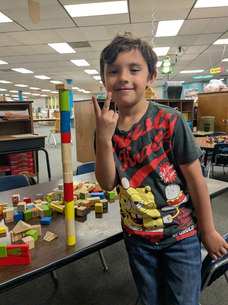 A child stands beside a table covered in colorful wooden blocks, proudly posing next to a tall block tower they built. They wear a SpongeBob-themed shirt, and the classroom background includes shelves, chairs, and learning materials.
