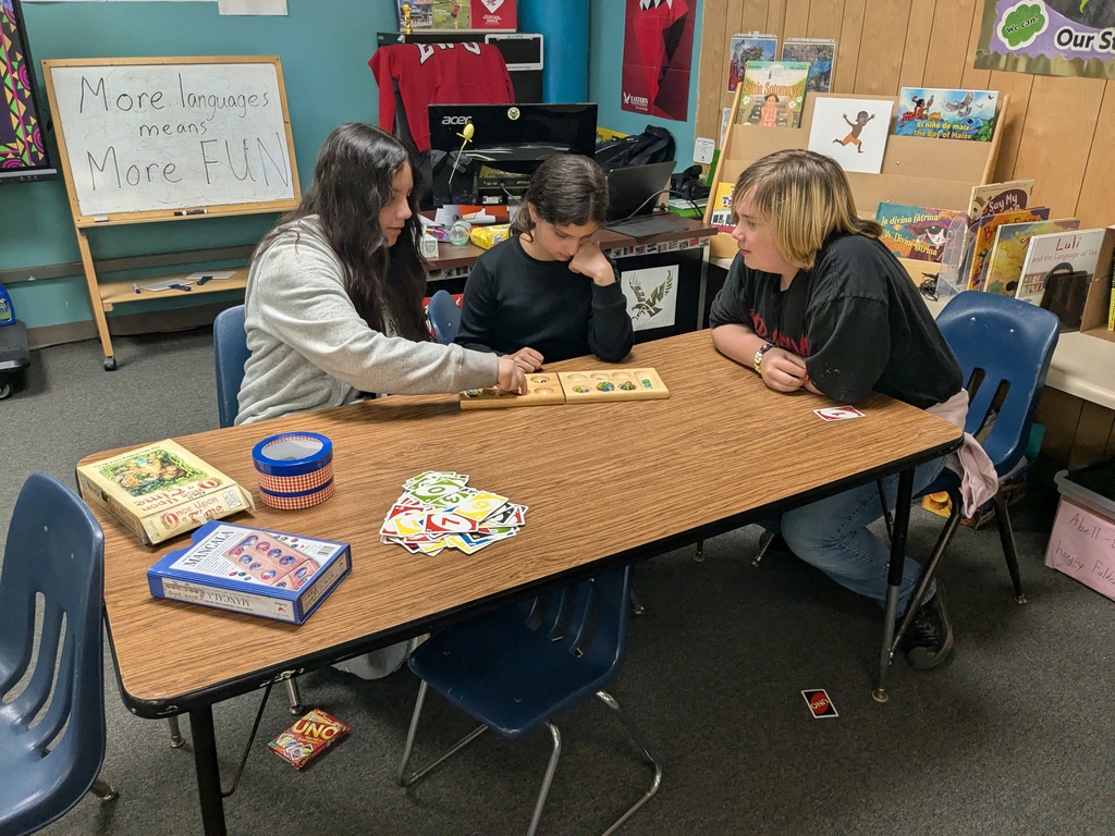 Three people sit around a classroom table playing Mancala, with UNO cards and other board games nearby. Behind them, a whiteboard reads “More languages means more fun,” and bookshelves and posters fill the colorful room.