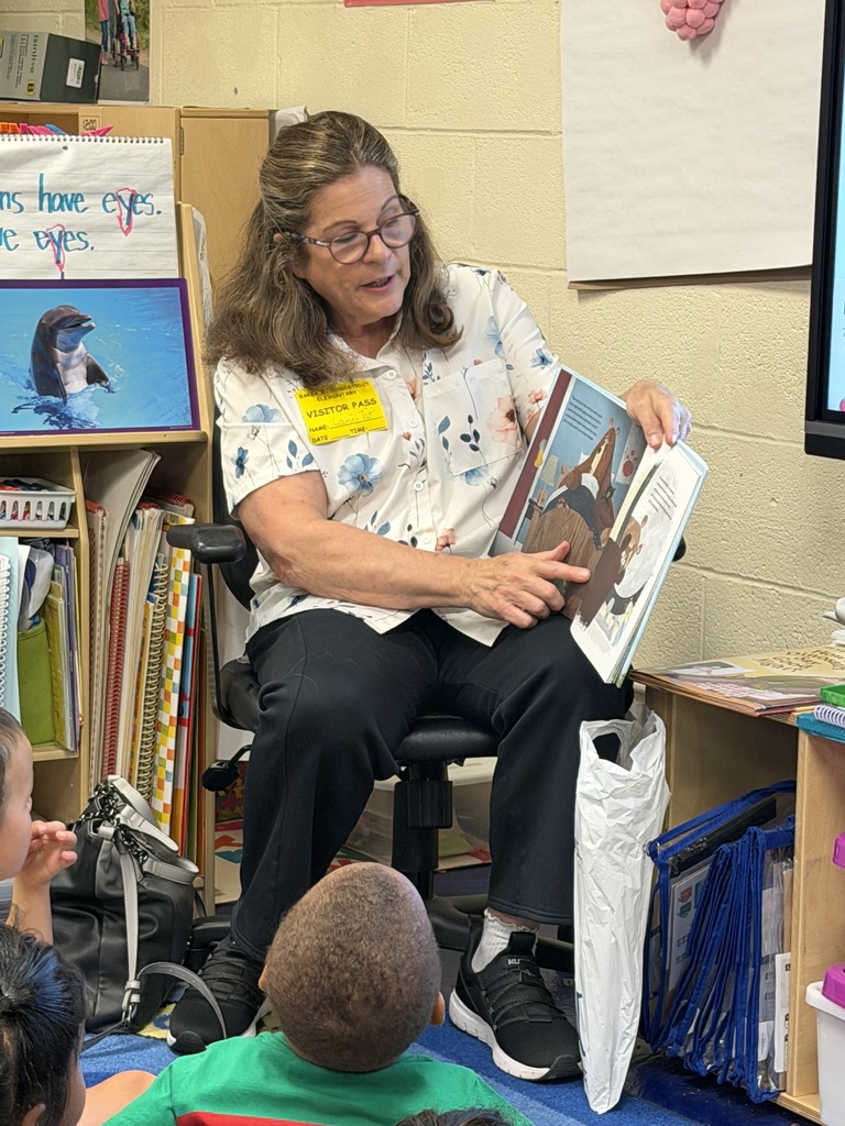 A close-up of Dr. Laurel Tait wearing a white floral shirt and a yellow visitor pass. She is smiling and pointing to an illustration of a bear while reading the book "I’m Not Scared, You’re Scared" to the students seated in front of her.
