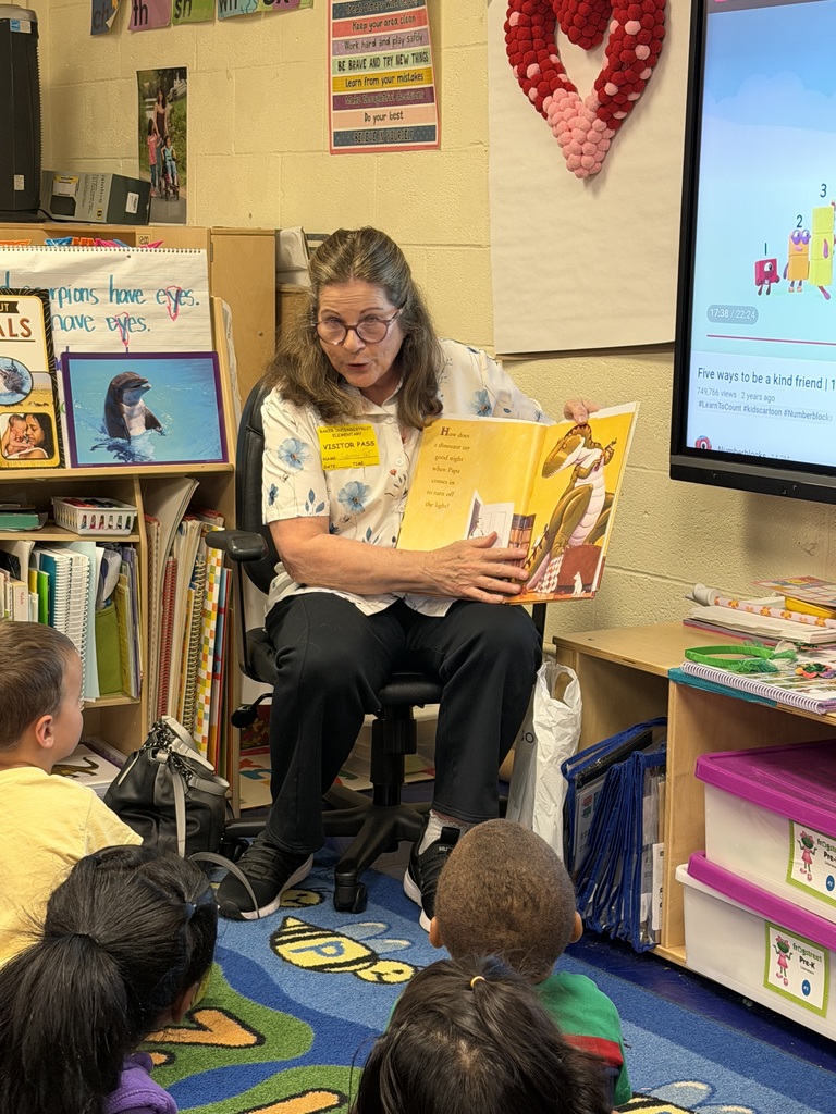Dr. Laurel Tait actively engages the Pre-K students while reading "How Do Dinosaurs Say Good Night?". She is holding the book open to show the children a colorful illustration of a large green dinosaur in a bedroom.