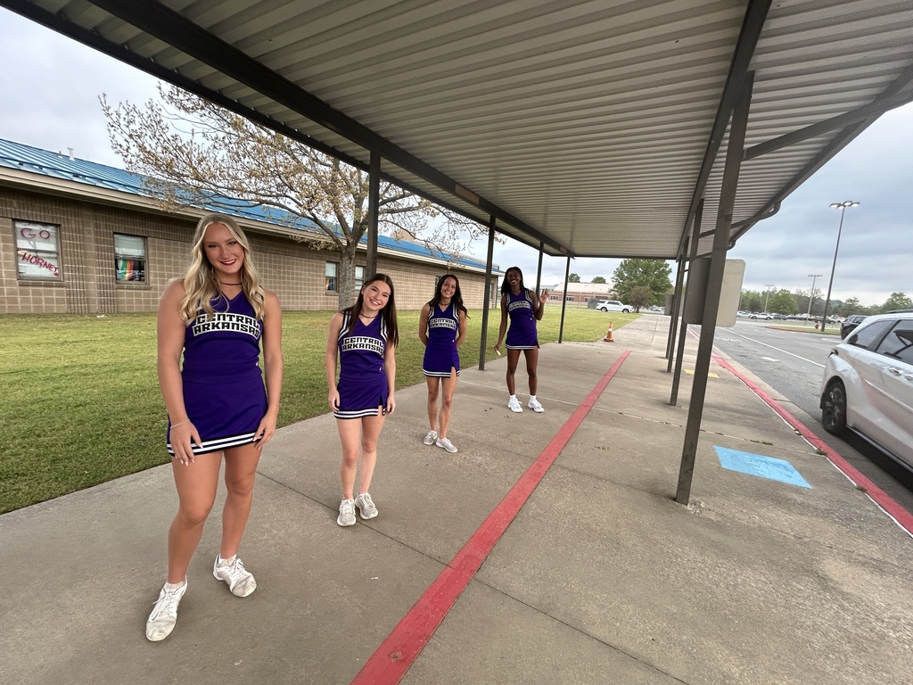 Four members of the University of Central Arkansas Cheer Squad stand under a covered walkway at Crystal Hill Elementary, posing in their purple uniforms before greeting arriving students.