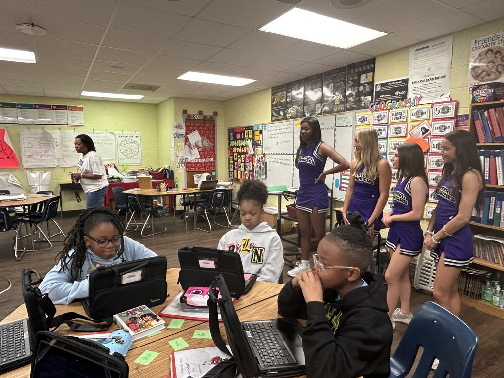 Students work at a round table on laptops while UCA cheerleaders visit the classroom, engaging with learners during their tour of Crystal Hill Elementary.