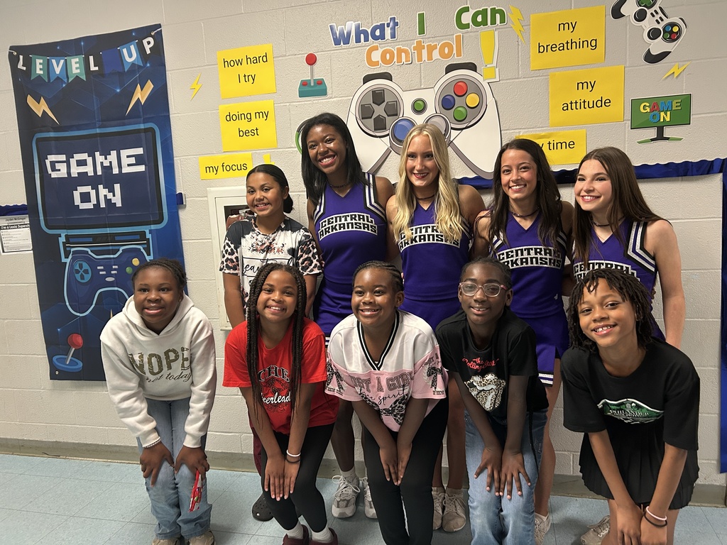 Four UCA cheerleaders stand with Crystal Hill Elementary students in a hallway decorated with gaming‑themed motivational posters during their classroom visits.