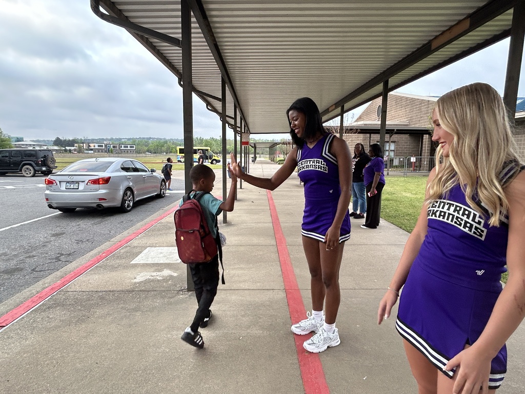 A UCA cheerleader gives a high‑five to a young student during morning arrival at Crystal Hill Elementary, with another cheerleader smiling nearby as cars line up behind them.