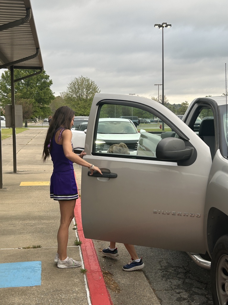 A UCA cheerleader in a purple uniform helps welcome students during morning drop‑off, opening a truck door for a student.