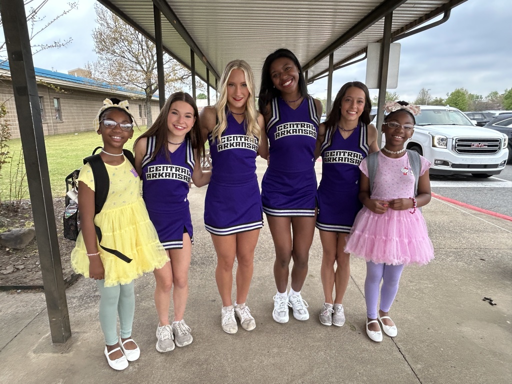 Three UCA cheerleaders pose with Crystal Hill Elementary students dressed in colorful outfits, all smiling under a covered walkway during the school visit.