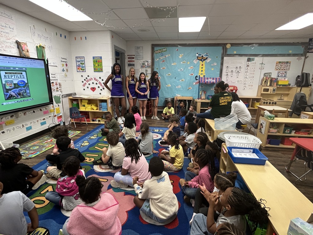 Young children sit on a colorful rug watching a reptile presentation while UCA cheerleaders stand at the front of the classroom, participating in Crystal Hill Elementary’s morning activities.