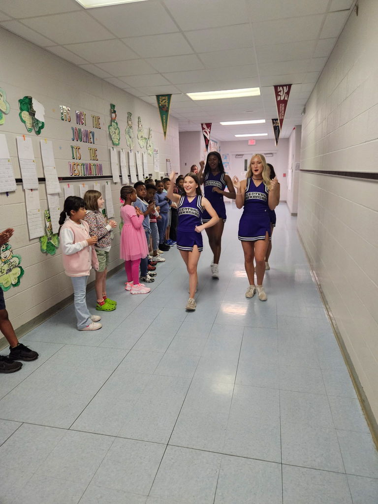 A line of Crystal Hill Elementary students stands along a hallway wall clapping as members of the University of Central Arkansas Cheer Squad walk past in their purple uniforms, creating a pep‑rally style welcome with student artwork and college pennants decorating the hallway.