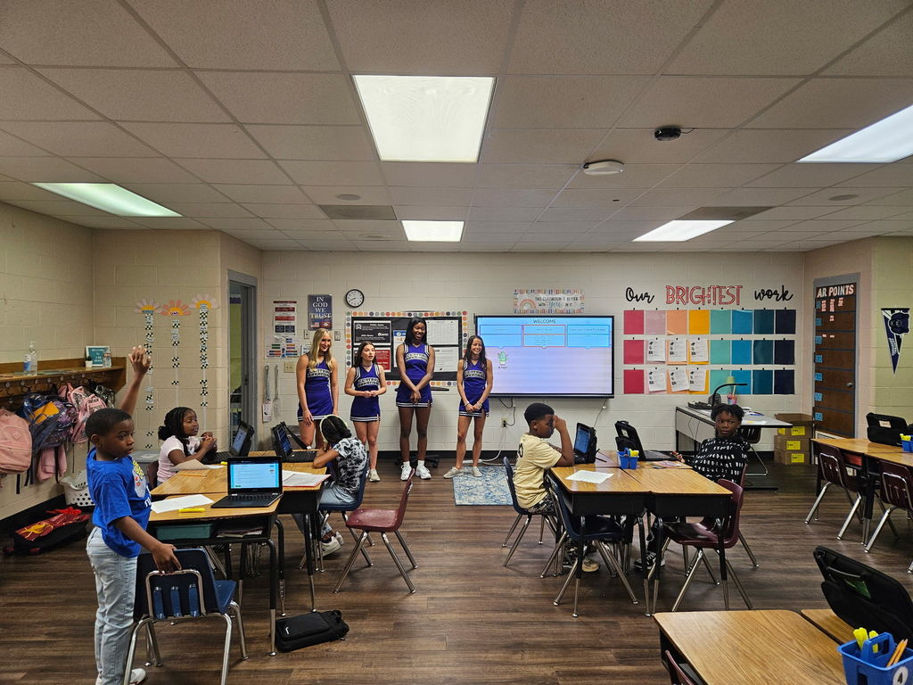 Four UCA cheerleaders stand at the front of a brightly decorated Crystal Hill Elementary classroom while students sit at grouped desks, one raising a hand, as a large screen displays a “Welcome” slide during the squad’s classroom visit.