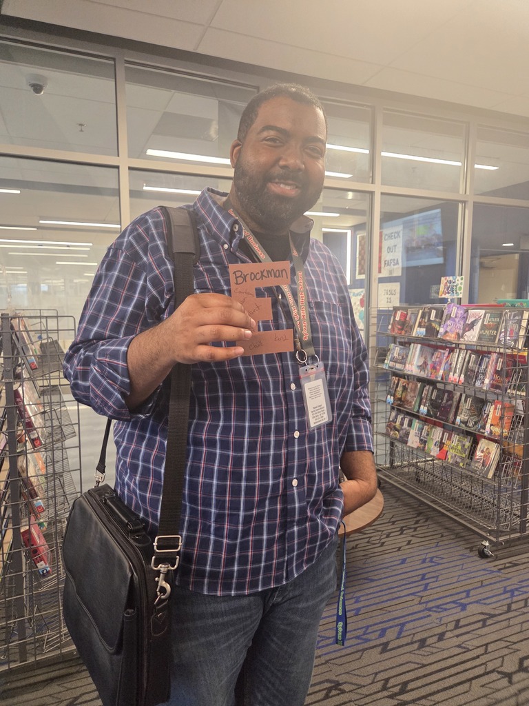 A Sylvan Hills High School teacher smiling while holding up a brown paper letter 'E', an April Fools' prank filled with positive messages from students.