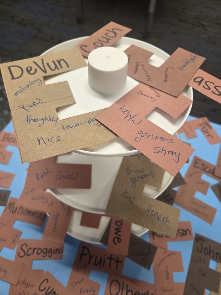 A close-up of a brown paper letter 'E' covered in handwritten compliments and positive notes from Sylvan Hills High School students to their teachers.