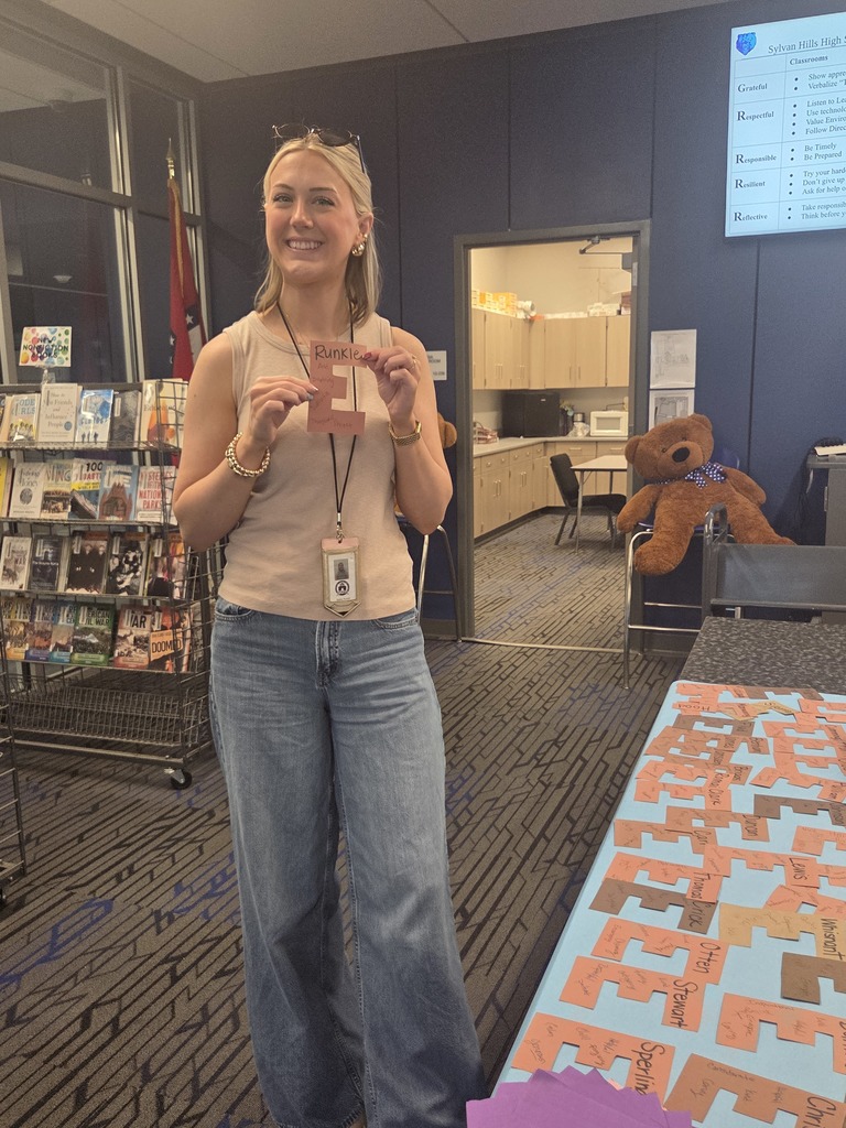 A Sylvan Hills High School teacher smiling while holding up a brown paper letter 'E', an April Fools' prank filled with positive messages from students.
