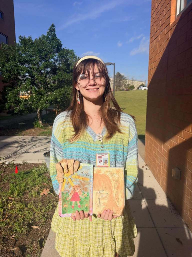 A student stands outdoors holding two mixed‑media art pieces, one a colorful landscape drawing and the other a textured abstract panel.