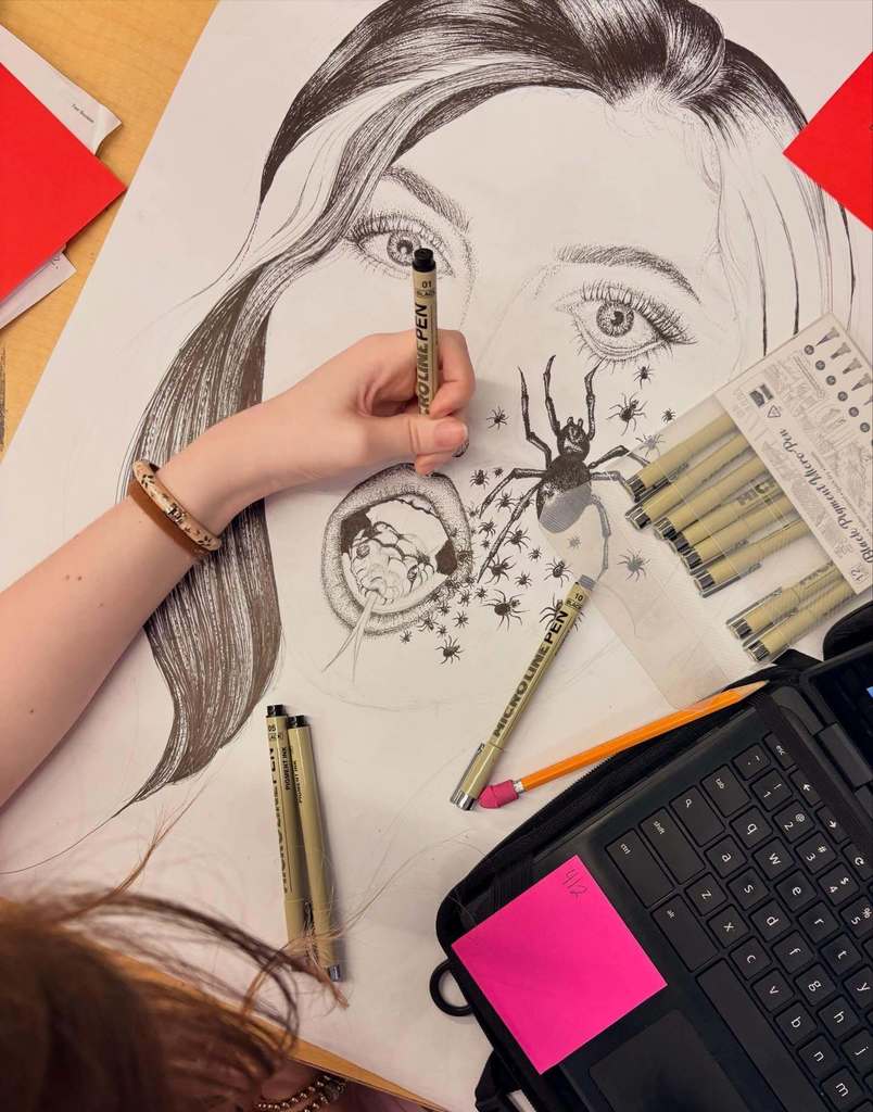 A student works on a detailed black‑and‑white pen drawing of a woman’s face with surreal spider imagery, using fine‑tip pens spread across the desk.