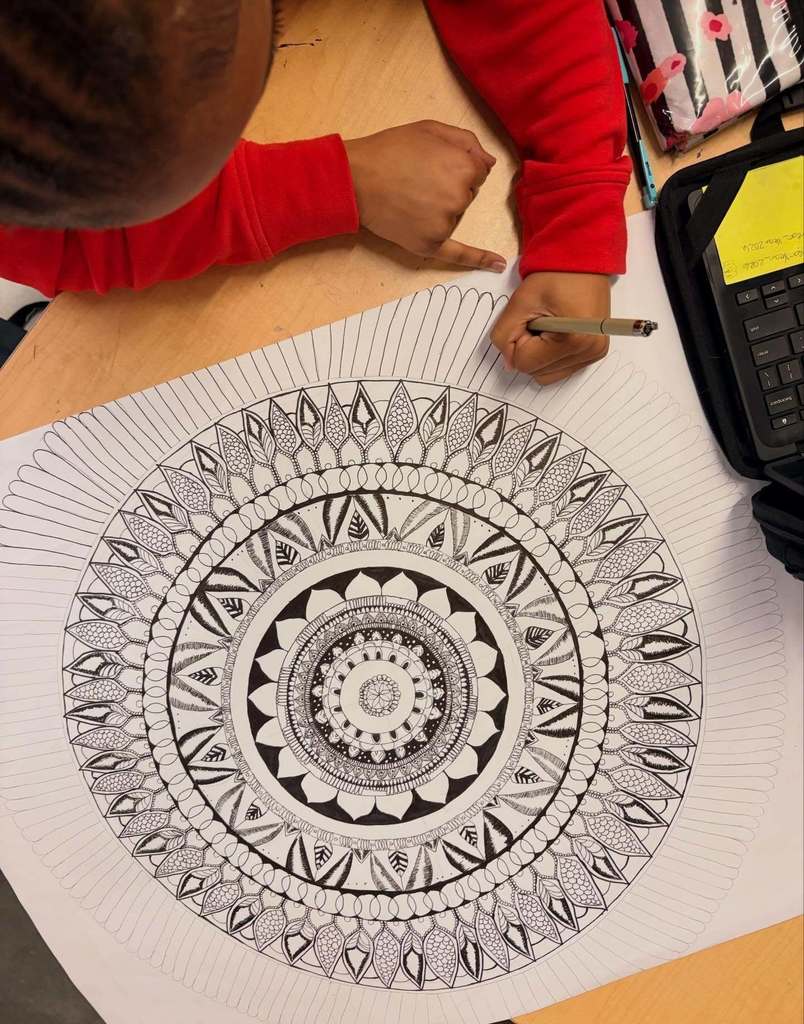 A student in a red shirt adds fine details to a large black‑and‑white mandala design on a table with a laptop and pencil case nearby.