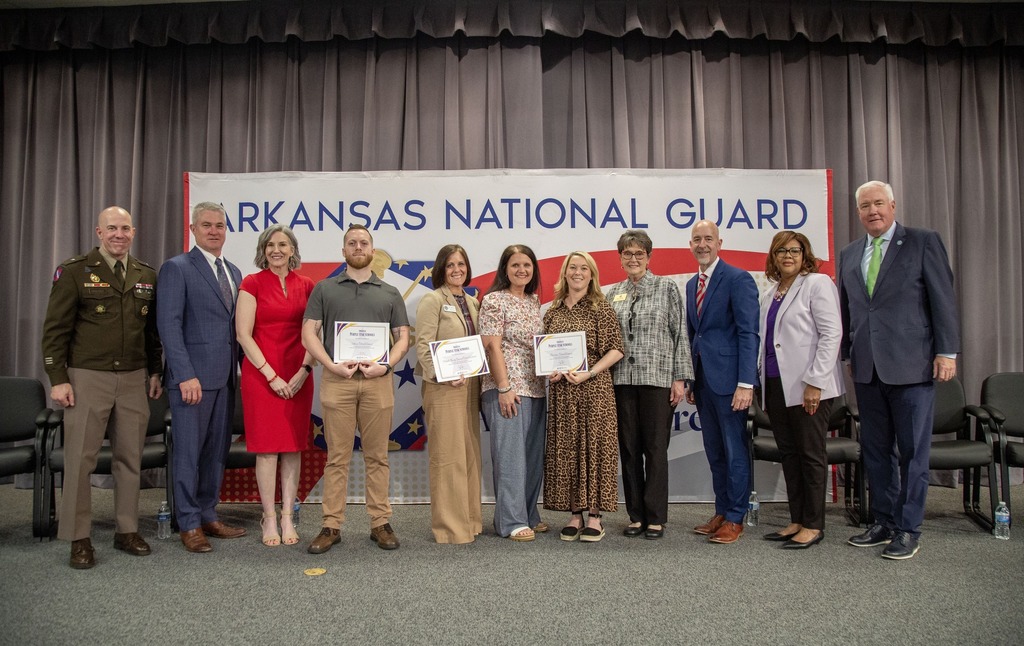 A group of ten people stand in front of an “Arkansas National Guard” banner, with three individuals in the center holding framed certificates during a recognition ceremony.