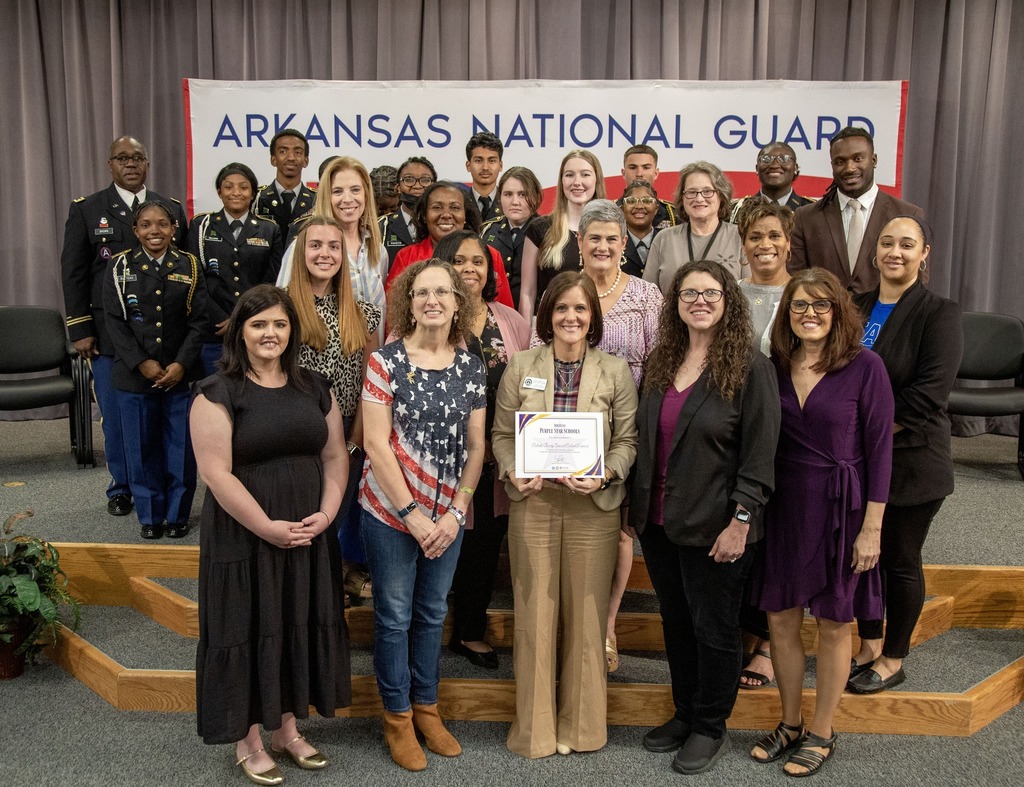 A large group of military personnel and civilians pose together on a stage in front of an “Arkansas National Guard” banner, with one person in the front row holding a framed certificate.