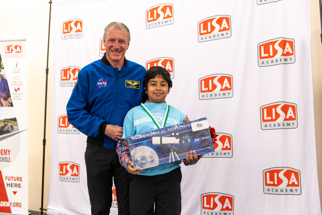 Two people stand in front of a backdrop covered with “LISA Academy” logos. On the left is retired NASA astronaut Dr. Michael Foale wearing a blue NASA jacket. On the right is 5th grader Shriyan Bhattacharrya holding a boxed Helium 80/600 astronomical telescope. Shriyan is wearing an Arkansas STEM Festival shirt and smiling after receiving his award.