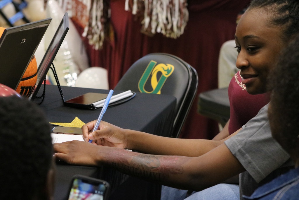 A student signs a document at a table with a basketball, phone, and framed item. The chair behind them displays a green and yellow Mills logo.