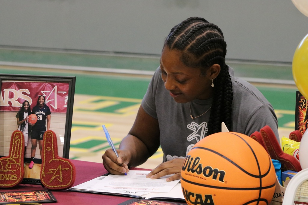 A student in a gray shirt signs a document at a maroon‑covered table decorated with a basketball, foam fingers, and a framed basketball photo.