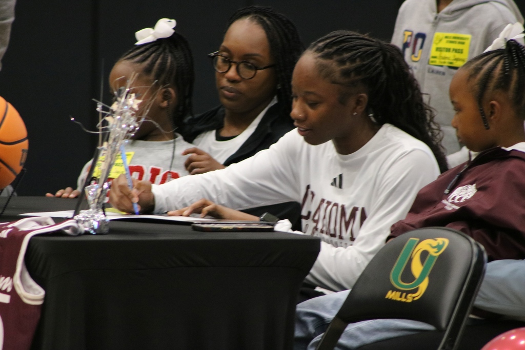 A student in a white shirt signs a document at a decorated table with a maroon jersey, basketball, and silver streamers. Three others, including two children, sit beside them.