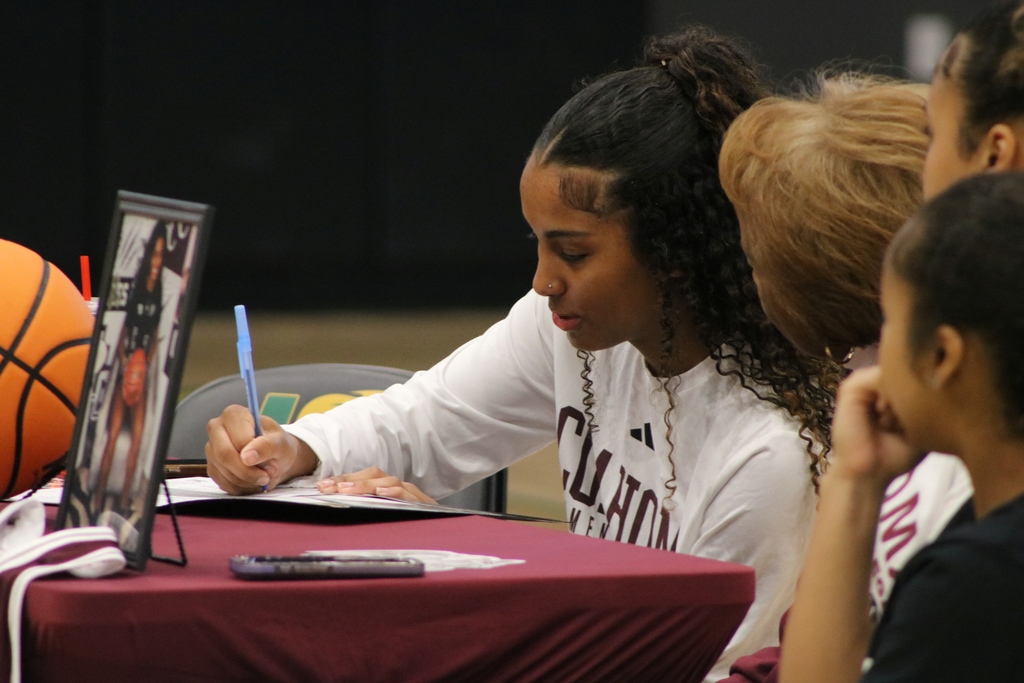 A student wearing a white long‑sleeve shirt signs a document at a maroon‑covered table with a basketball and framed photo. Two people sit nearby watching.
