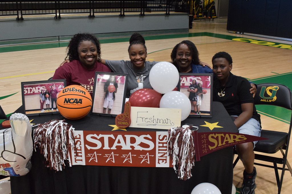 Four people sit at a decorated table on a basketball court with balloons, framed photos, pom‑poms, and an Oklahoma City University pennant. A banner reads “These Stars Shine.”