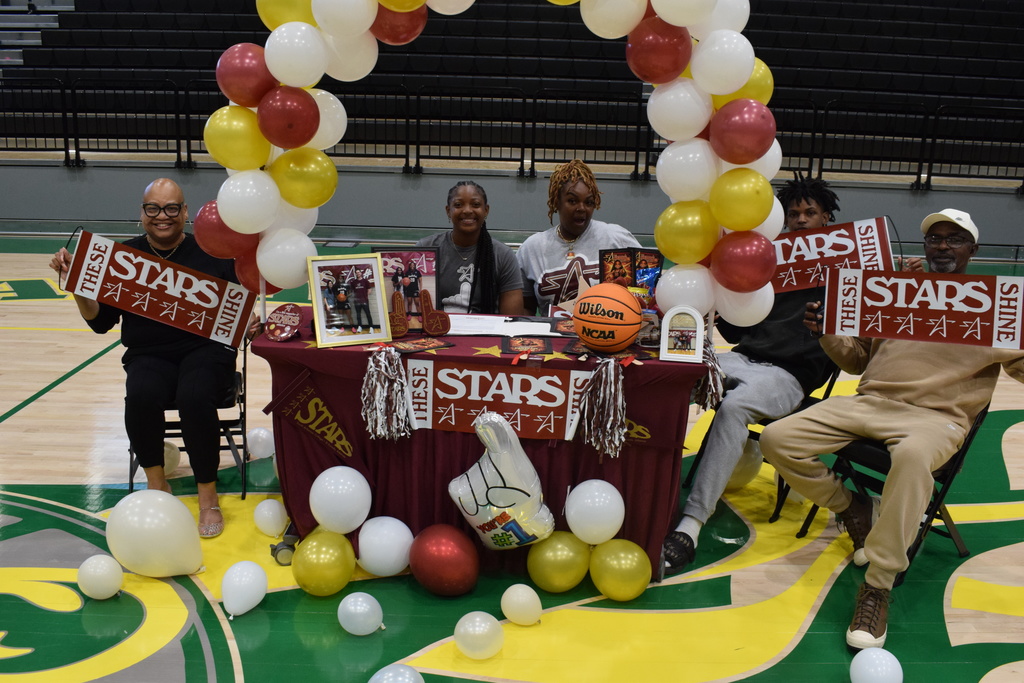 Five people sit behind a maroon table decorated with balloons, pom‑poms, framed photos, and a basketball. They hold signs reading “These Stars Shine” during a signing ceremony.