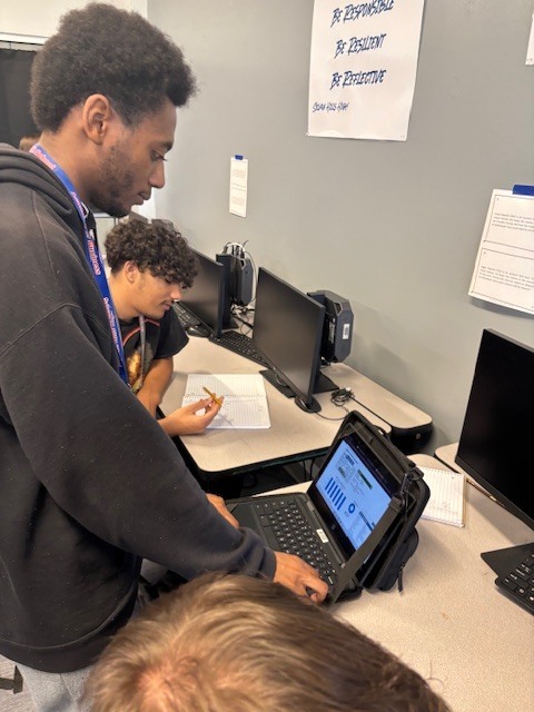 Three students collaborate at a desk with computer monitors and a tablet displaying charts and graphs. One writes in a notebook while another points at the tablet. A classroom sign on the wall reads “Be Responsible, Be Respectful, Show Integrity.”