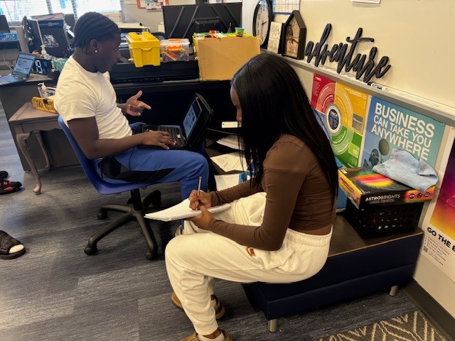 Two students work in a classroom. One sits at a desk using a laptop and gesturing, while the other writes in a notebook. The space is decorated with posters and supplies, including a sign that says “BUSINESS CAN TAKE YOU ANYWHERE.”