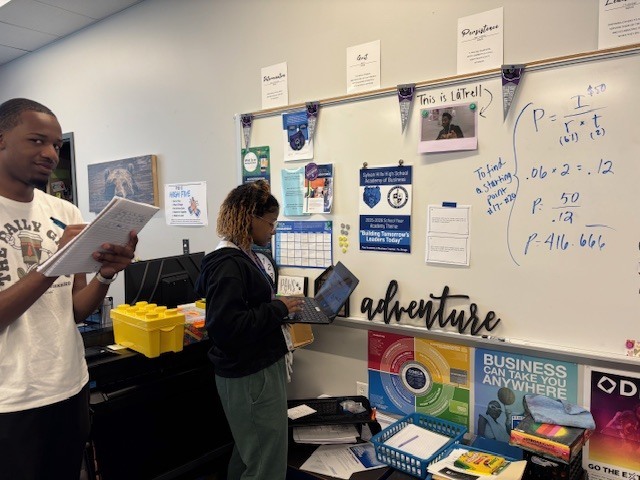 Two students stand in a classroom. One holds a notebook and pen, and the other works on a laptop near a whiteboard covered in handwritten math notes and formulas. The wall behind them has posters, bins, and classroom materials.
