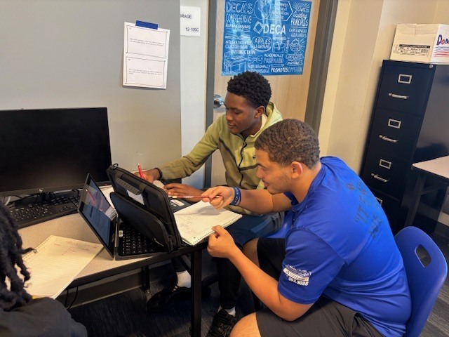 Two students sit at a desk working together. One writes in a notebook while the other points at a laptop screen. Papers, notebooks, and a desktop computer are on the desk, with a DECA poster visible on the wall behind them.
