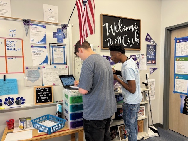 Two students stand at a small classroom workstation with a laptop and storage drawers. The wall behind them is decorated with posters, schedules, and a letter board that says “DAILY CHECK-IN HOW AM I FEELING TODAY?” An American flag hangs above the area.