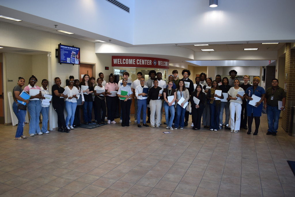 A large group poses for a photo in front of a Welcome Center counter. Many hold papers or folders, and a digital screen behind them displays an app advertisement.