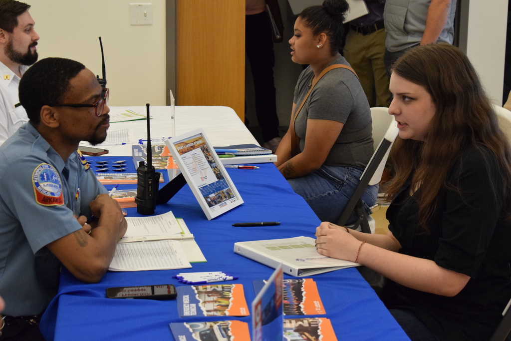 A MEMS representative in a light blue uniform talks with a student at a table displaying brochures, pens, and a framed information sheet about student and career pathways.